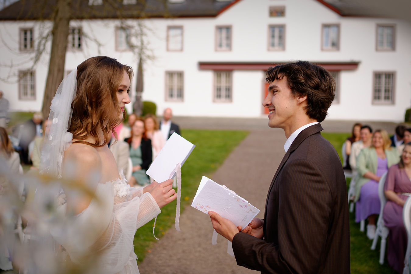 Braut und Bräutigam halten sich an den Händen und lächeln sich während ihrer freien Trauung vor einem weißen Gutshaus an.